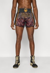 Μαύρα και κόκκινα σορτς Venum με περίπλοκα σχέδια, ελαστική μέση και χρυσές λεπτομέρειες. Το μοντέλο φοράει μαύρα αθλητικά παπούτσια.