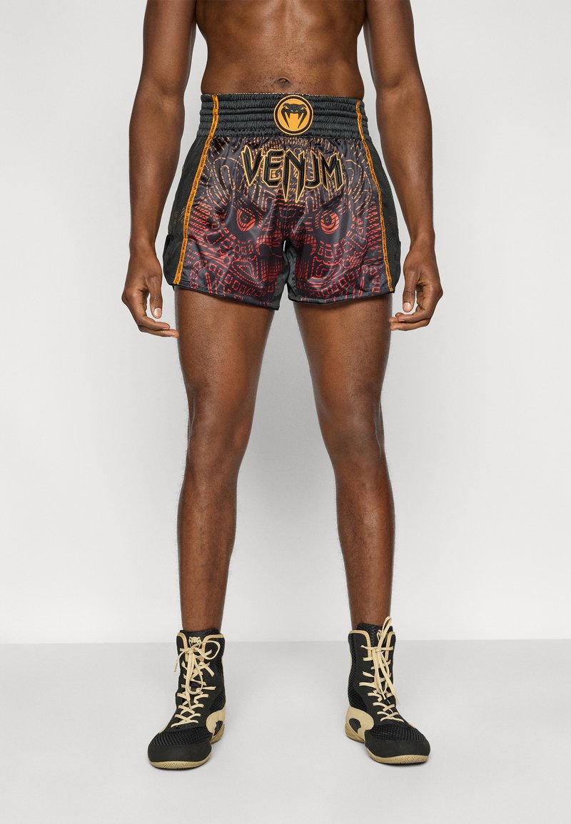 Μαύρα και κόκκινα σορτς Venum με περίπλοκα σχέδια, ελαστική μέση και χρυσές λεπτομέρειες. Το μοντέλο φοράει μαύρα αθλητικά παπούτσια.