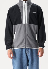 Fleecejacka med hög krage, i svart och grå färgblock, vit dragkedja och sidofickor. Mjuk textur med elastiska ärmslut.