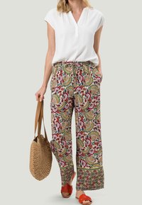 Haut blanc à manches courtes, pantalon large à motifs fleuris dans des tons de rouge, vert et or. Accessoires : sac tissé et sandales rouges.