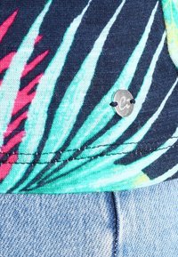 Koszula z tkaniny w tropikalny wzór z motywami roślinnymi w kolorach zielonym, niebieskim i różowym; mała metalowa zawieszka z literą "C"; gładka faktura.