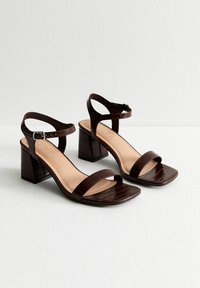 Un par de sandalias de tacón bloque en color marrón oscuro con grabado de cocodrilo, con tiras en el tobillo y diseño de punta cuadrada abierta sobre una superficie blanca.