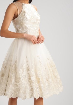 Femme portant une robe blanche sans manches longueur genou avec une superposition en dentelle florale dorée et un col transparent, mains délicatement jointes à la taille.