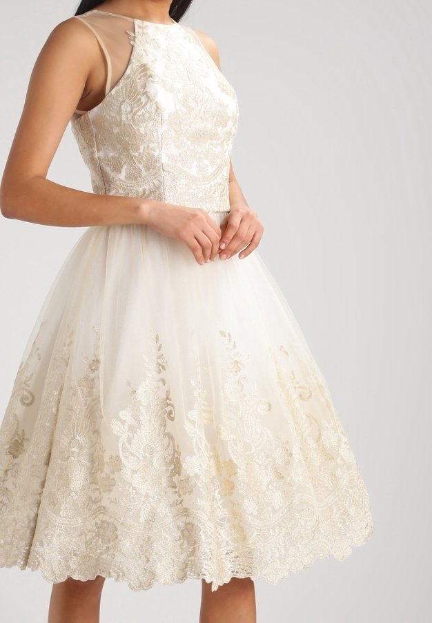 Femme portant une robe blanche sans manches longueur genou avec une superposition en dentelle florale dorée et un col transparent, mains délicatement jointes à la taille.