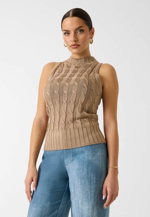 Femme portant un haut beige sans manches en maille avec des motifs torsadés et un jean denim bleu clair, debout devant un fond uni.
