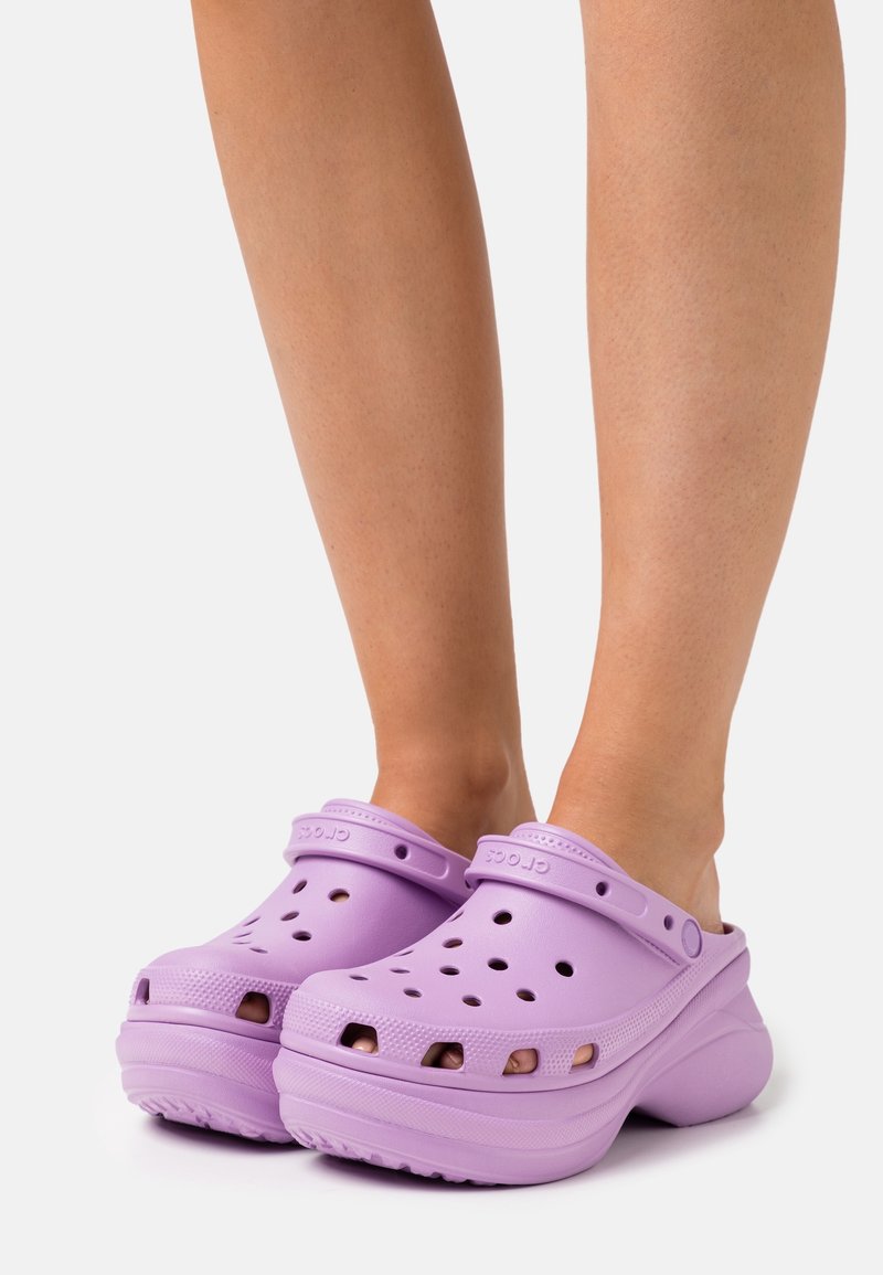 Jambes chaussées de Crocs plateformes lavande avec des trous perforés et des brides de talon ajustables sur un fond clair uni.
