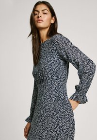 Das marineblaue Blumenmaxikleid hat einen runden Ausschnitt, lange Ärmel mit rüschigen Manschetten und eine figurbetonte Silhouette, die mit kleinen weißen Blumen verziert ist.