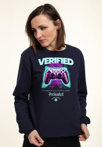 Marineblaues Sweatshirt mit langen Ärmeln. Zentrales Grafikdesign zeigt einen lila Gamecontroller und den Text "VERIFIED GAMER" in lebendigen Farben.