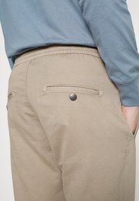 Beige broek met een gladde textuur, voorzien van twee achterzakken met knoopsluitingen en een elastische tailleband. Draag met een lichtblauw langsliepend shirt.