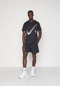 Μαύρο κοντομάνικο μπλουζάκι Nike με λευκό λογότυπο, συνδυασμένο με μαύρο σορτς και λευκά αθλητικά παπούτσια, τοποθετημένο σε ουδέτερο φόντο.