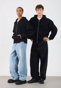 Sudaderas con cremallera negras llevadas por dos personas, combinadas con jeans holgados—uno de un azul claro y otro de un azul oscuro—destacando un ajuste relajado y estilos contrastantes.