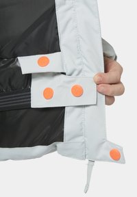 Světle šedá podšívka bundy s oranžovými cvoky, černou podšívkou a kapsou na zip. Disponuje hladkým povrchem a zesíleným šitím.