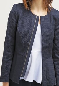 Granatowa teksturowana kurtka z pikowanym wzorem, z metalowym przednim suwakiem i baskinką. Nosi się ją na białą bluzkę.