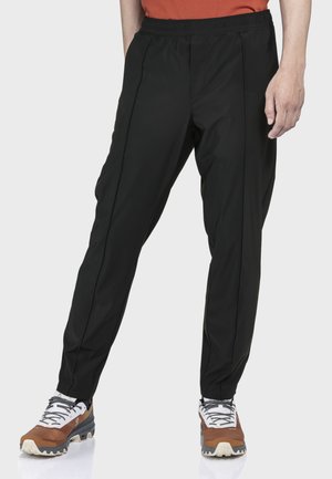 Mann trägt schwarze, schmal zulaufende Hosen und braun-weiße Wanderschuhe, steht vor einem einfachen hellen Hintergrund.