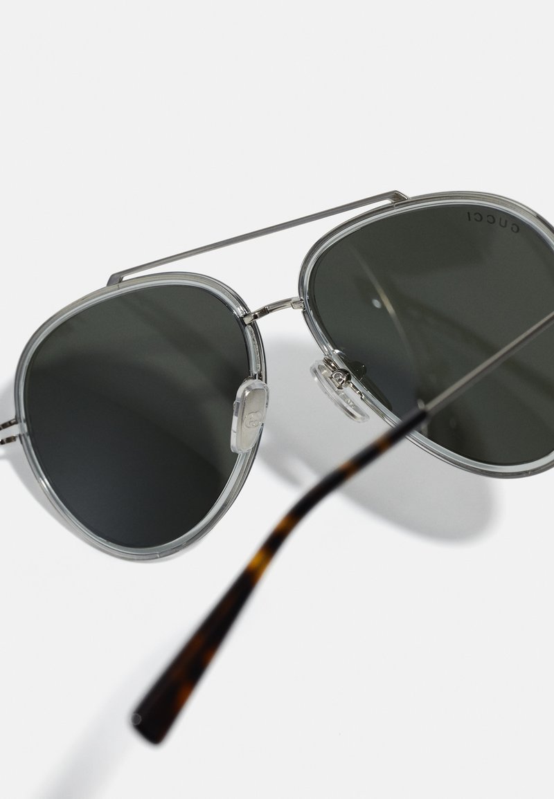 Lunettes de soleil à monture en métal argenté avec des verres gris foncé, arborant une forme ovale, des accents transparents et des extrémités de branches en écaille.