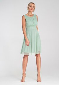 Miętowo-zielona dopasowana sukienka z plisowanym gorsetem i wąskim paskiem w talii. Spodnica do kolan w luźnym kroju z przezroczystą nakładką oraz bez rękawów.