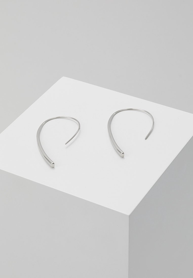 Boucles d'oreilles créoles en argent au design minimaliste, forme allongée, texture lisse et structure légèrement incurvée, présentées sur un socle blanc.