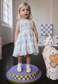 Biele šaty s modrými mašľami a čiernymi bodkami, kombinované s bielymi sandálmi. Stojí na kruhovej fialovej platforme vedľa maľovanej sochy mačky.