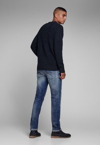 Marineblauwe gebreide trui met een gestructureerd kabelpatroon, gecombineerd met lichtblauwe jeans en donkere laarzen, die een casual stijl van achteren laat zien.