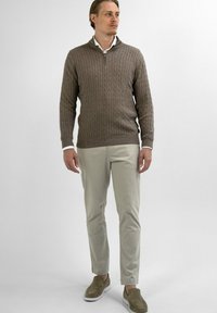 Bruine kabelgebreide trui met een ritssluiting in de kraag, gecombineerd met lichtbeige chino's en taupe instappers, op een effen achtergrond.