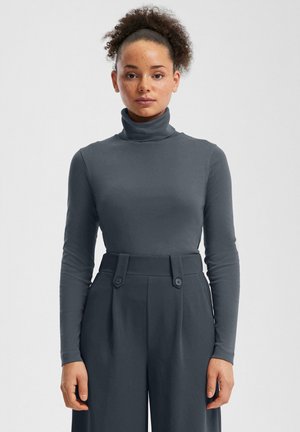 Siva turtleneck majica dugih rukava, u kombinaciji s visokim sivo-hlačama širokih nogavica s dvostrukim gumbima. Glatka tekstura kroz cijelu odjeću.