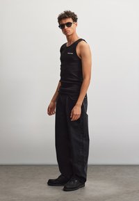 Man met kort, krullend haar die een zwarte zonnebril, een aansluitend zwart hemd, losse donkere jeans en zwarte schoenen draagt, staat tegen een effen achtergrond.