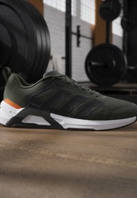Chaussure de sport vert foncé et noire avec semelle blanche posée sur le sol d'une salle de sport avec des disques d'haltérophilie et du matériel en arrière-plan.