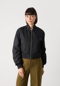 Giacca bomber nera accorciata con una texture liscia e lucida, maniche a costine e cinturino in vita, dotata di una zip frontale e tasche laterali.
