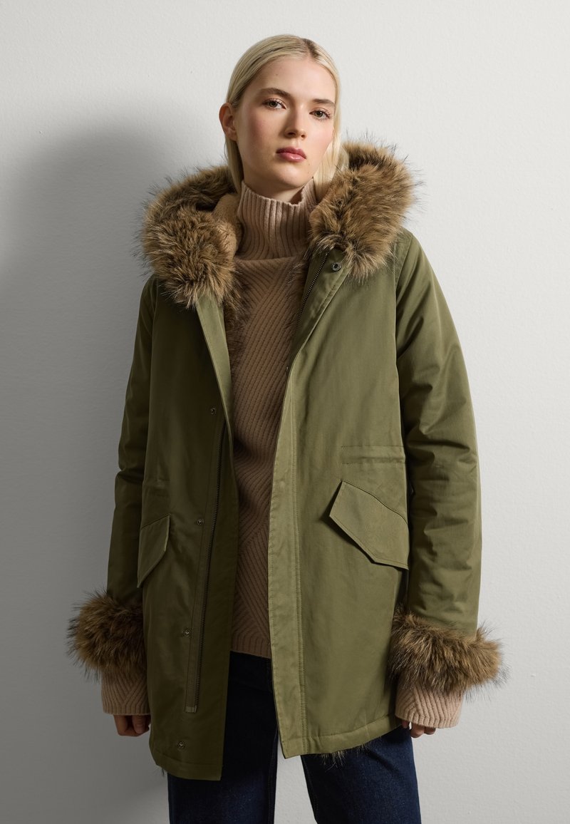 Parka verde oliva con borde de piel sintética en la capucha y los puños, con cremallera en el frente, dos bolsillos frontales y un suéter de punto en color marrón debajo.