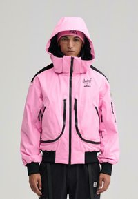 Mand iført en knaldrød pink hættetrøje med sorte lynlåsedetaljer og matchende pink hue, stående mod en enkel lys baggrund.