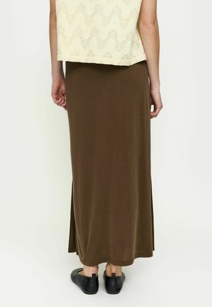 Personne portant une longue jupe marron avec des fentes latérales, un haut sans manches couleur crème avec un motif en dentelle, et des chaussures noires sans lacets, debout de dos.