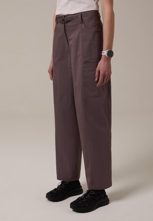 Persona vistiendo pantalones sueltos marrón oscuro con un bolsillo lateral, zapatillas negras y un reloj inteligente blanco en la muñeca izquierda.