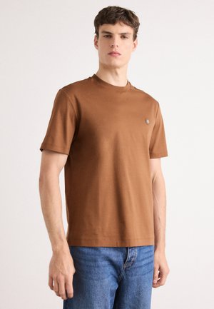 T-shirt marron à manches courtes en coton doux, avec un col rond et un petit logo rond sur la poitrine gauche. Porté avec un jean bleu.
