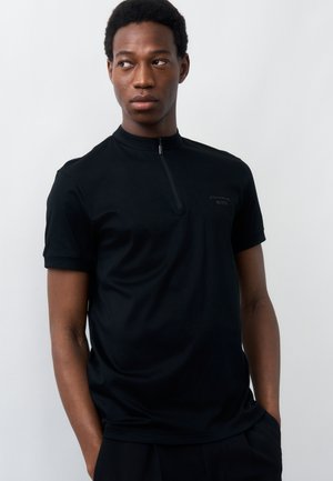 Νεαρός άντρας με κοντά μαλλιά που φοράει μαύρο κοντομάνικο πουκάμισο με φερμουάρ, κοιτάζει ελαφρώς προς τα αριστερά μπροστά σε απλό φωτεινό φόντο.