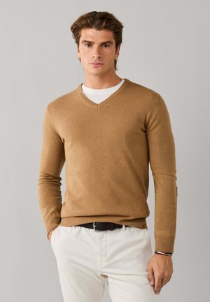 Hombre de pie con un suéter marrón de cuello en V sobre camisa blanca, pantalones blancos, cinturón negro y botas oscuras con cordones, sobre un fondo liso.