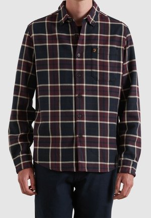 Hombre con una camisa de botones de cuadros azul marino y burdeos con mangas largas y un bolsillo en el pecho con un logo bordado.