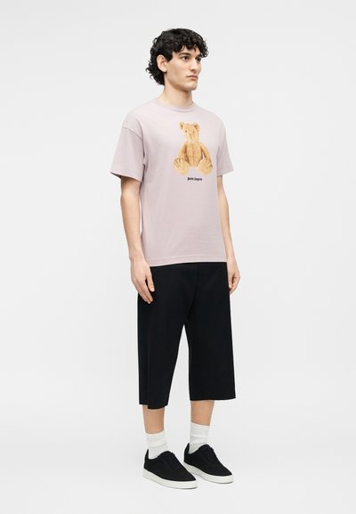 Νεαρός άνδρας που φοράει ανοιχτό μοβ μπλούζα με στάμπα αρκουδάκι, μαύρο παντελόνι με φαρδιά μπατζάκια μέχρι τον αστράγαλο, λευκές κάλτσες και μαύρα αθλητικά παπούτσια, όρθιος πλαγίως.