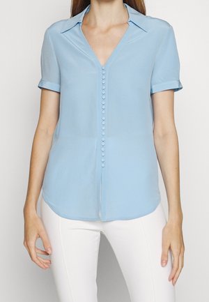 Femme portant un chemisier bleu clair à manches courtes avec des boutons cachés sur le devant et un pantalon blanc, vue du cou aux hanches.