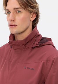 Giacca con cappuccio bordeaux con colletto zip, dotata di una fodera in rete e del logo VAUDE sul petto. Materiale liscio e leggero.