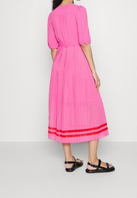 Rosa plissierte Midi-Kleid mit kurzen Puffärmeln und einem rot gestreiften Saum; kombiniert mit schwarzen Riemchensandalen. Glatte Stofftextur.