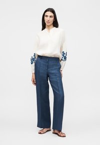 Femme debout portant un chemisier blanc aux manches à fleurs bleues, un pantalon bleu taille haute à jambes larges et des sandales marron.