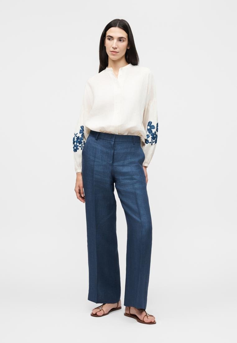 Femme debout portant un chemisier blanc aux manches à fleurs bleues, un pantalon bleu taille haute à jambes larges et des sandales marron.