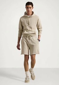 Νεαρός άνδρας περπατάει, φορώντας μπεζ φούτερ, ταιριαστά σορτς, λευκές κάλτσες και μπεζ αθλητικά παπούτσια, σε έναν απλό ανοιχτό φόντο.