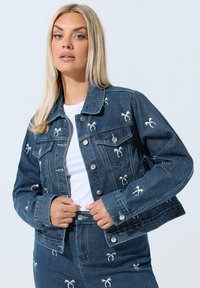 Denimtakki, jossa on vaaleansiniset brodeeratut nauhat, klassinen kaulus, etutaskut ja hopeanväriset napit. Yhdistetty valkoisen paidan kanssa.