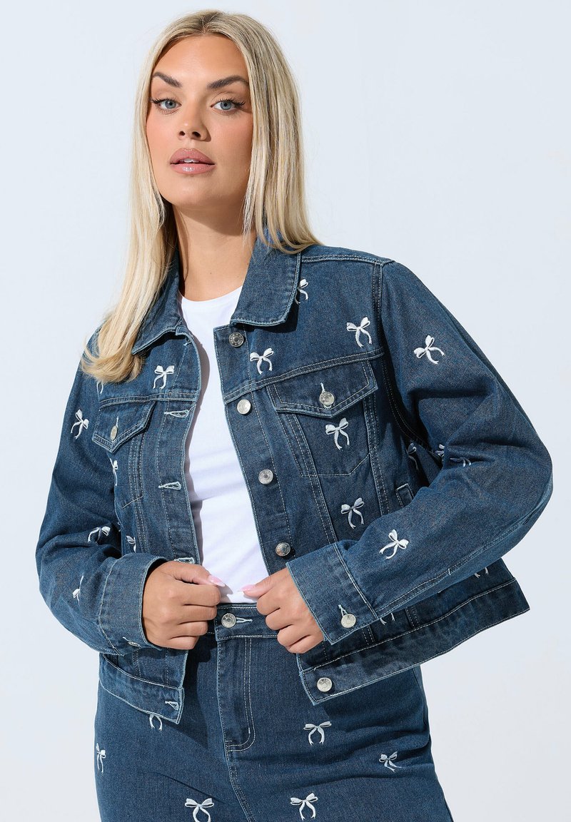 Denimtakki, jossa on vaaleansiniset brodeeratut nauhat, klassinen kaulus, etutaskut ja hopeanväriset napit. Yhdistetty valkoisen paidan kanssa.