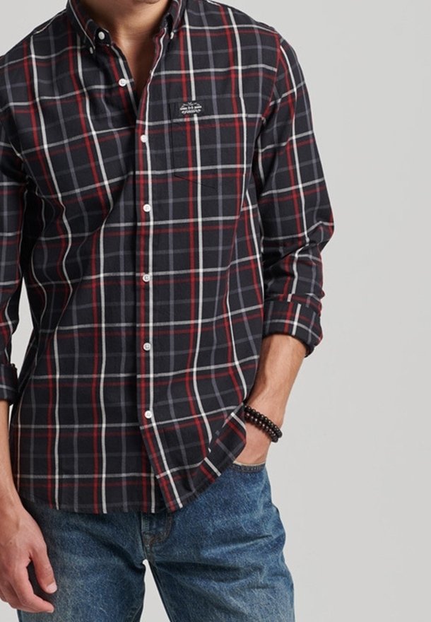 Camisa de cuadros en negro y rojo con botones, mangas largas, bolsillo en el pecho y botones blancos, usada sobre pantalones vaqueros azules.