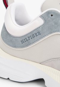 Zapatilla gris claro con parte superior de malla blanca, acentos de ante en azul claro, suela de goma y "HILFIGER" grabado en la parte posterior.