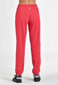 Röda joggare med resårmidja som smalnar av mot anklarna, visade från baksidan. Svarta sneakers med mönstrade remmar är synliga.