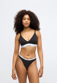 Crna bralette s podesivim naramenicama i bijelim Calvin Klein logotipom na rubu; usklađeni crni tange gaćice sličnim logotipom na rubu.