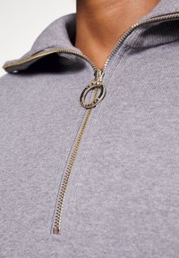 Maglione grigio con zip, caratterizzato da una texture a coste, con zipper dorata e linguetta circolare. Dettagli ravvicinati del colletto e della zip.